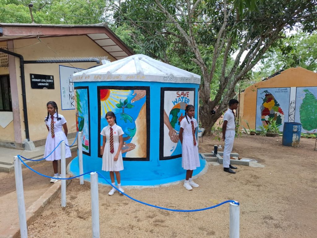 Schoolgirls standing near and protecting a rainwater harvesting tank at their blue school in Sri Lanka