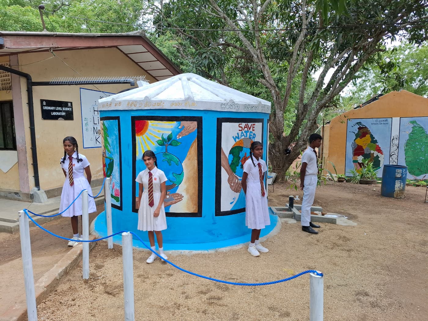 Schoolgirls standing near and protecting a rainwater harvesting tank at their blue school in Sri Lanka