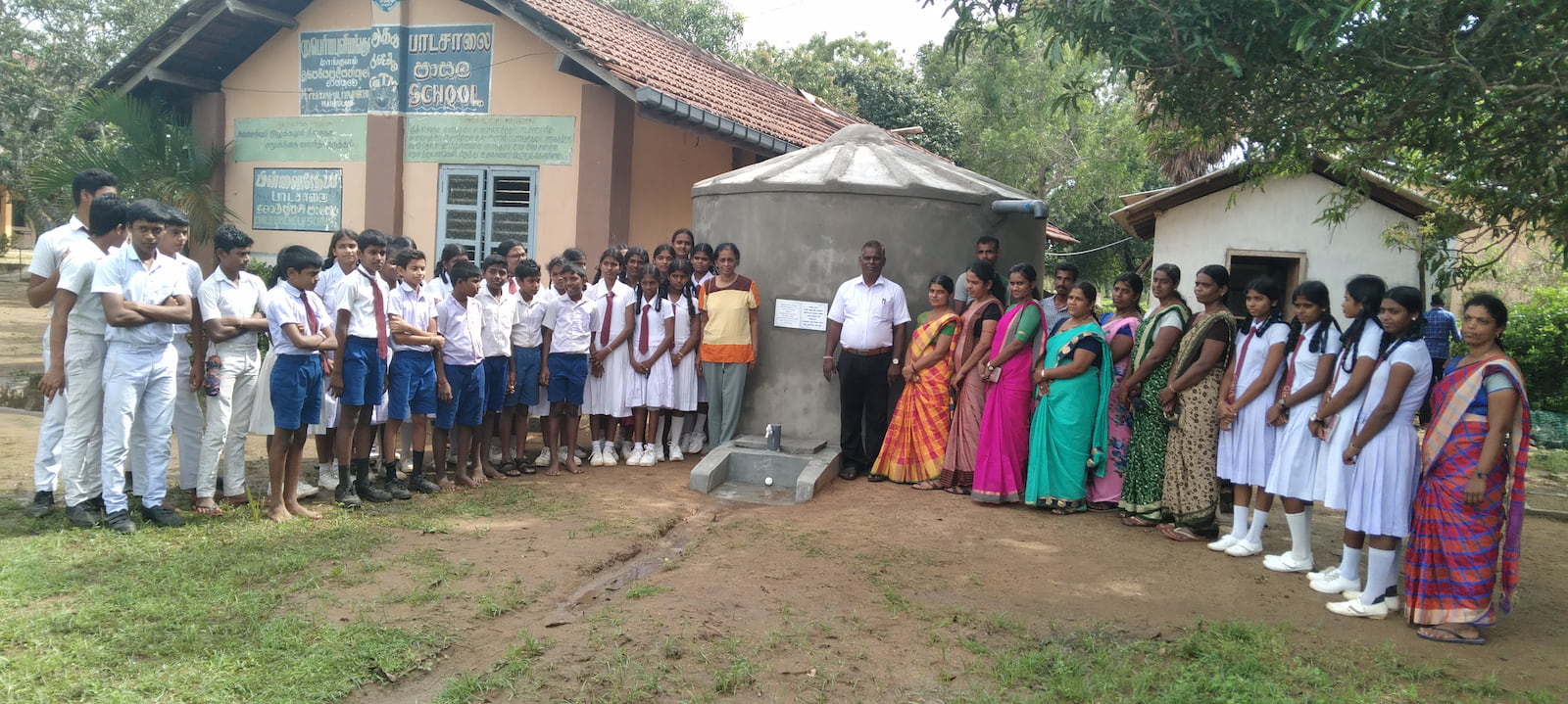 Blue school staff and children standing together in front of a rainwater harvesting tank in Sri Lanka